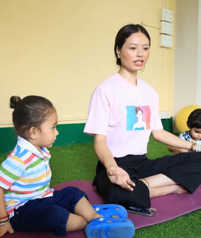 A teacher leading two toddlers through a seated yoga pose