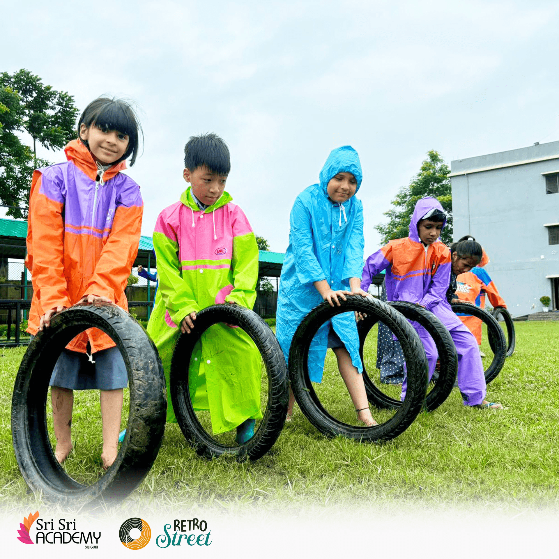 Sri Sri Academy students rolling tyres through an outdoor obstacle activity in raincoats